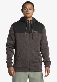 Men's zip-up hoodie in dark brown and black, featuring a textured fabric and a drawstring hood, with front pockets and a logo patch.