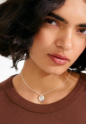 Silberne Halskette mit einem runden Anhänger, der ein Rosenmotiv zeigt, getragen zu einem braunen Hemd. Der Anhänger ist fein gearbeitet und glänzt.