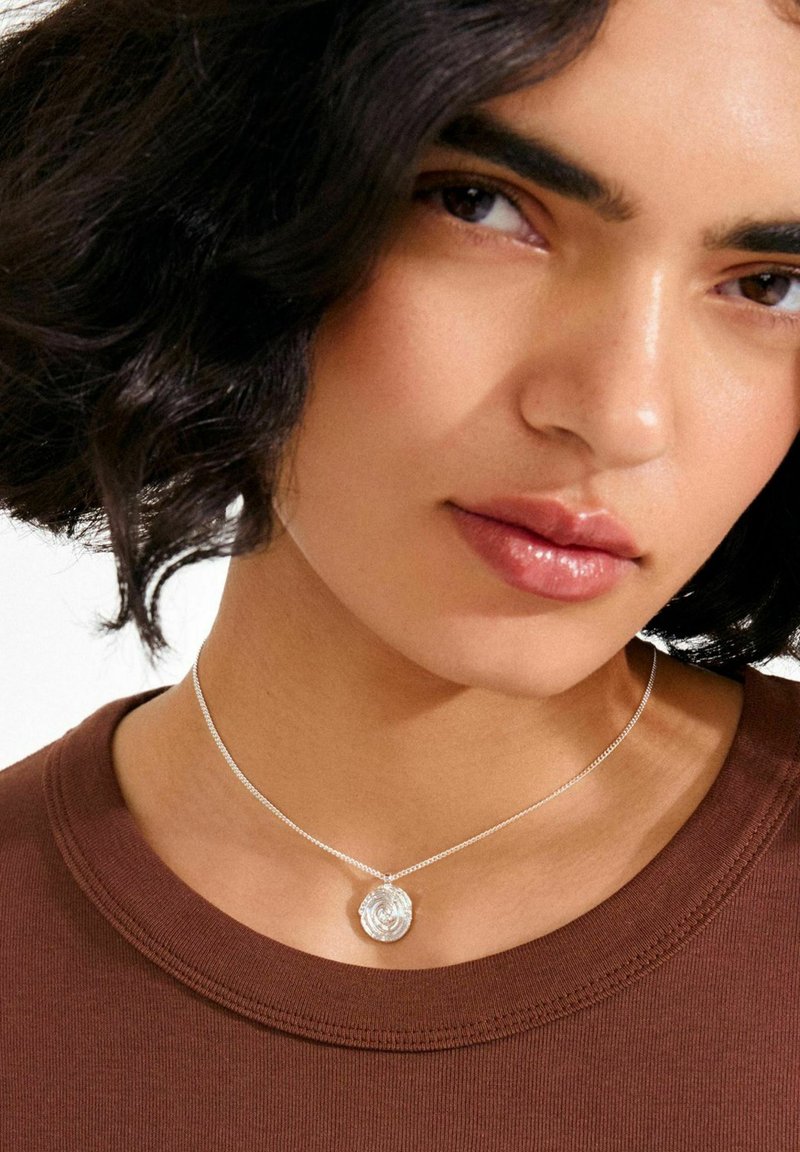 Collar de plata con un colgante circular con diseño de rosa, llevado sobre una camisa marrón. El colgante tiene detalles intrincados y un acabado brillante.