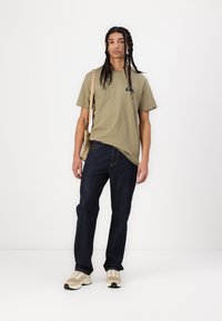Olivengrønn bomull T-skjorte med en liten logo, kombinert med mørkeblå denimjeans og beige sneakers. Modell som holder en lys veske.