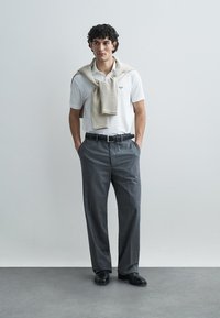 Polo branca, calças cinzentas com padrão, suéter bege pousado sobre os ombros, cinto preto e sapatos pretos. Modelo em pé com as mãos nos bolsos.