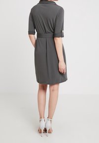 Šedé šaty s krátkými rukávy a pásem v pase, vyrobené z hladké tkaniny. Mají límečkový výstřih a dekorativní knoflíky na manžetách.