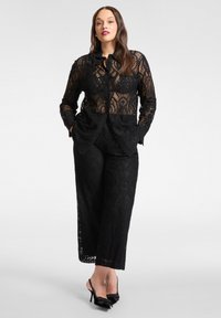 Czarny koronkowy strój składający się z przejrzystej koszuli zapinanej na guziki oraz dopasowanych spodni z misternymi wzorami. Dopełniony czarnymi butami na wysokim obcasie.
