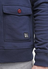 Felpa blu navy con un tessuto testurizzato, tasca frontale con un bottone rosso e un piccolo etichetta del marchio all'orlo.