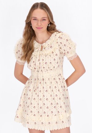 Ung kvinde iført en cremefarvet kjole med ballonærmer og rødt blomstermønster, stående med hænderne bag ryggen mod en hvid baggrund.