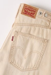 Beige denim jeans met een gestructureerde afwerking, voorzien van een bruine leren patch met rode tekst, en een achterzak met het iconische Levi's stikselontwerp.