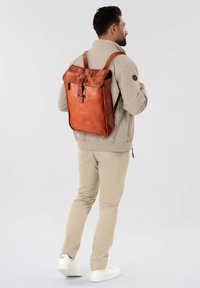 Zaino in pelle marrone con pattina pieghevole, dotato di chiusura a fibbia e spallacci regolabili, indossato da una persona.