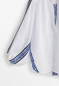 Blouse blanche à manches longues avec broderie géométrique bleue le long des manches, des côtés et de l'encolure, présentant une coupe ample et une petite ouverture en V.