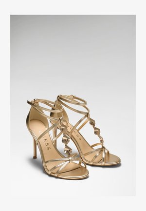 Paar goldene Sandalen mit hohen Absätzen, Knöchelriemen und dekorativen, gedrehten Metallakzenten an den vorderen Riemen.