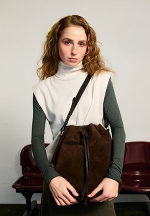 Woman with curly red hair wearing a white sleeveless turtleneck over a green long-sleeve top, holding a large brown suede bucket bag.