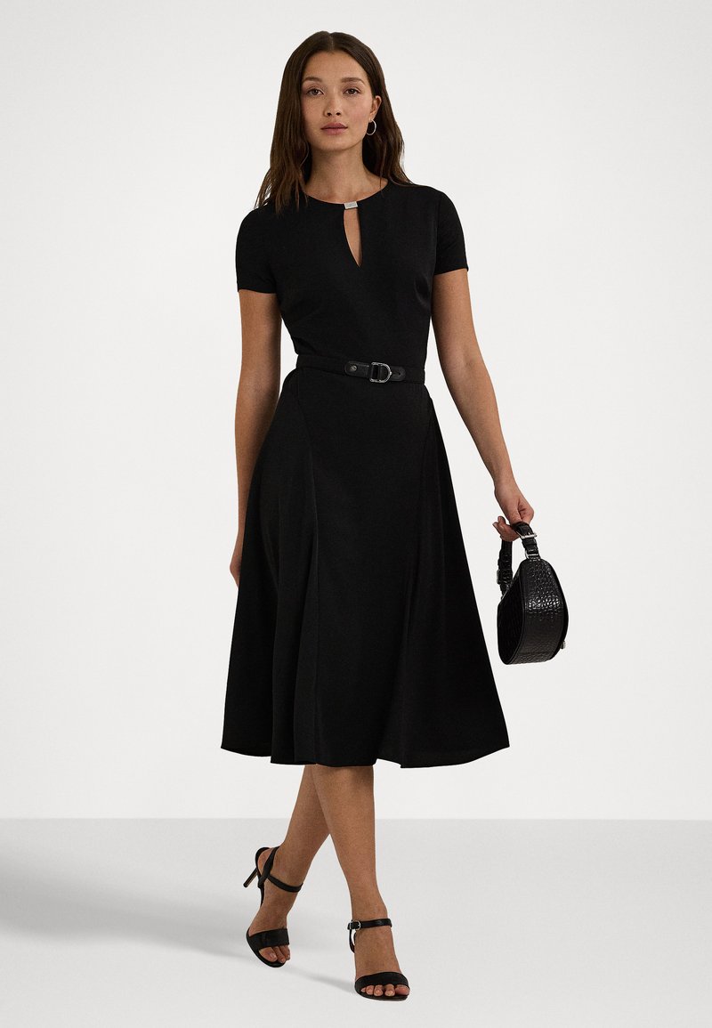 Abito nero con maniche corte, scollatura a chiave, vita cintrata e gonna fluida. Abbinato a décolleté nere con cinturini e una borsa in tessuto.