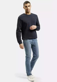 Granatowy sweter z długim rękawem, okrągłym dekoltem i subtelnym logo. Połączony z jasnoniebieskimi dżinsami i białymi trampkami, stojący na neutralnym tle.