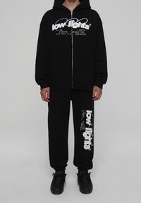 Svart zip-up hoodie och matchande byxor i mjukt material, med vita och blå logografik på båda plaggen och en avslappnad passform.