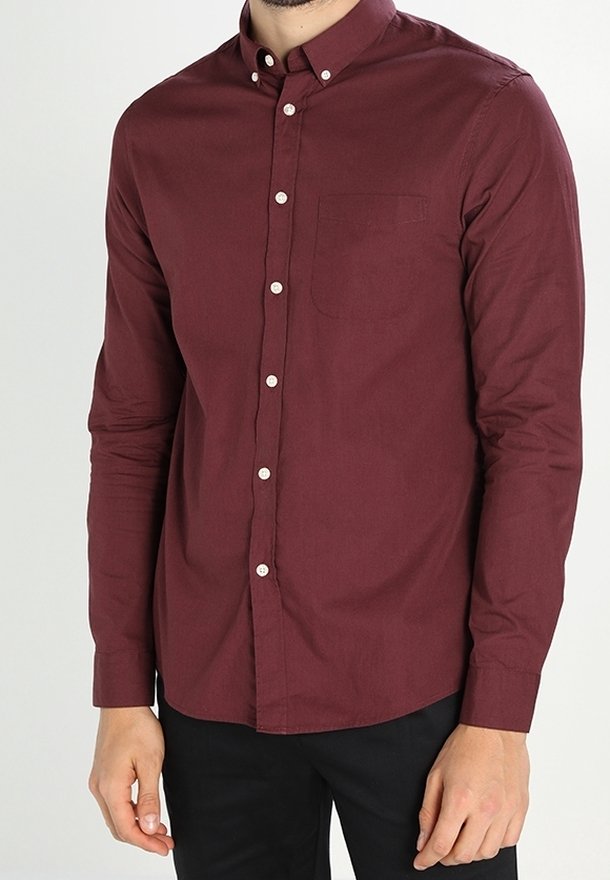 Un homme portant une chemise bordeaux à manches longues, boutonnée avec une poche poitrine, associée à un pantalon noir, se tient devant un fond neutre.