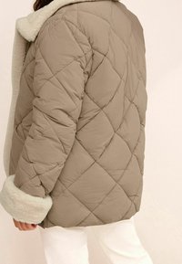Chaqueta acolchada beige con un acogedor cuello y puños de piel sintética crema, que presenta un diseño de costuras en forma de diamante y un corte holgado.