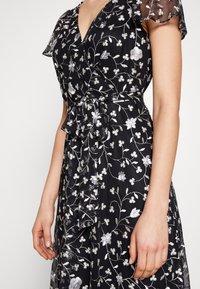 Černé šaty s květinovou výšivkou v stříbrné a bílé barvě, krátkými volánovými rukávy, výstřihem do V a zavazovacími pásky v pase. Měkká, průsvitná textura.