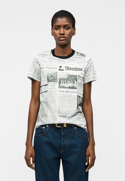 Femme portant un t-shirt à manches courtes imprimé de journaux avec un col noir et un jean bleu, sur un fond clair uni.