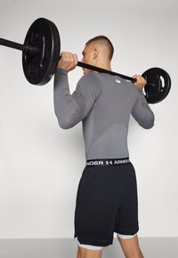 Graues langärmliges Sportoberteil mit passformgerechtem Design und gesticktem Logo, kombiniert mit schwarzen Shorts mit einem weißen, gebrandeten Bund, während er eine Langhantel hält.