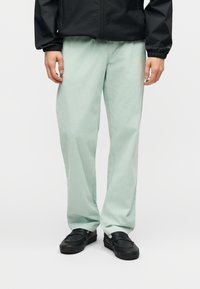 Άτομο που φοράει ανοιχτό πράσινο παντελόνι άνετης εφαρμογής, μαύρα παπούτσια χωρίς κορδόνια και μαύρο μπουφάν με ελαστικές μανσέτες, στέκεται μπροστά σε απλό φόντο.