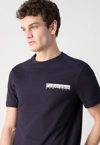 Tamsiai mėlynas medvilninis marškinėliai trumpomis rankovėmis ir apvaliu kaklu; kairėje krūtinės pusėje yra baltas Calvin Klein logotipas.