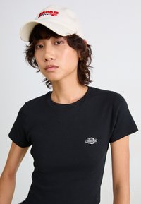 Sort bomulds t-shirt med korte ærmer og et lille hvidt Dickies-logo. Beige kasket med rødt broderet tekst "Racing." Glat stoftekstur.