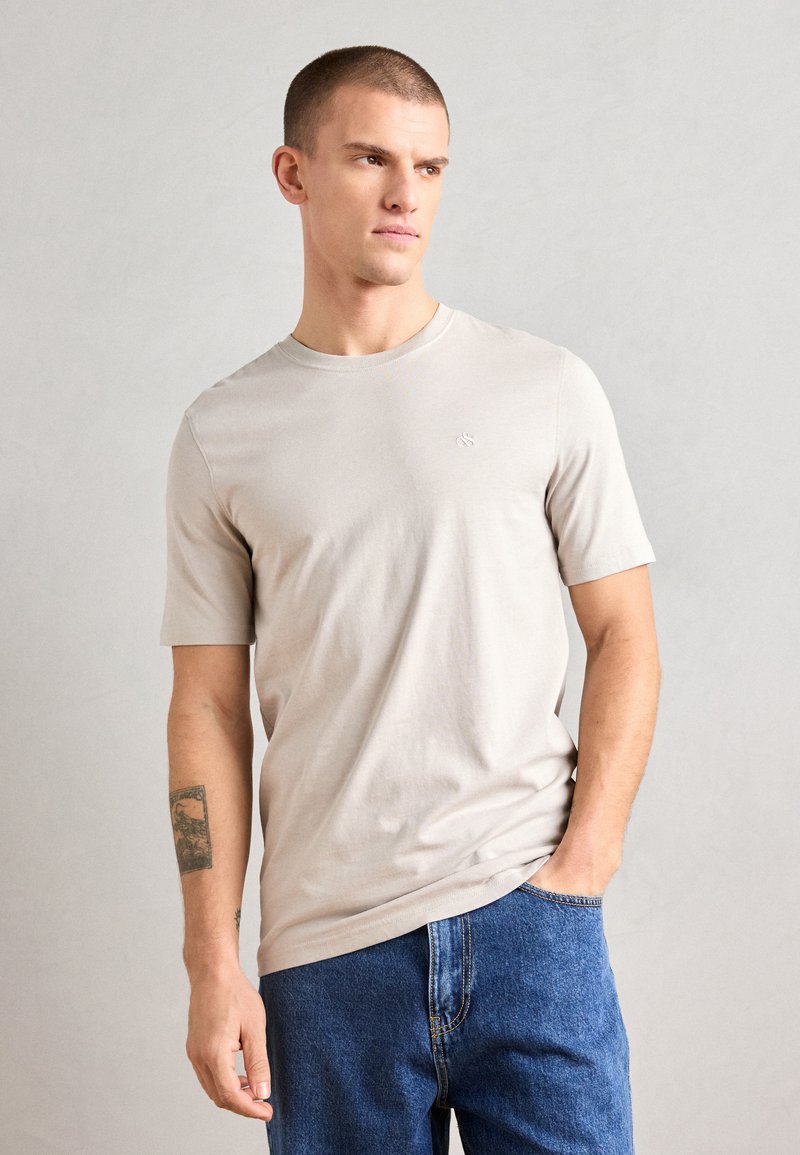 Beige kortermet t-skjorte i mykt stoff med rund hals. Har en liten logo på brystet. Kombinert med blå denimjeans.