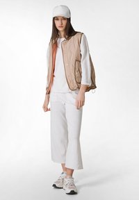 Beige gesteppte Weste mit Druckknöpfen, kombiniert mit einem weißen Langarmshirt und hellgrauen lockeren Hosen. Weiße Sneaker runden den Look ab.