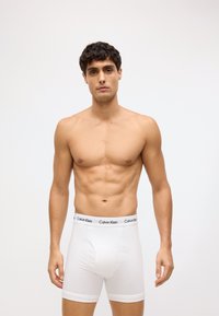 Calvin Klein bijele bokserice s crnim logotipom na pojasu. Tkanina izgleda glatko s prilagođenim dizajnom i blagim rastezanjem.