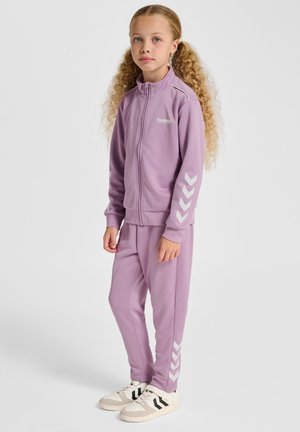 Paarse trainingspakset bestaande uit een jas met rits en een wit logo en chevronaccenten, gecombineerd met bijpassende broek. De schoenen zijn beige met zwarte strepen.