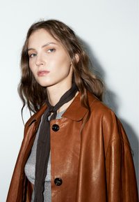Bruin leren jas met een ronde kraag en een oversized silhouet, gecombineerd met een donkere stofsjaal over een grijze top, tegen een witte achtergrond.