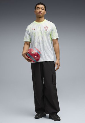 Młody mężczyzna trzymający czerwoną i niebieską piłkę nożną, ubrany w białą koszulkę reprezentacji Portugalii i czarne spodnie cargo na szarym tle.