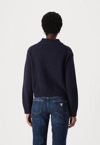 Pull court bleu marine avec un col côtelé haut et un bas côtelé. Porté avec un jean bleu foncé présentant un logo triangulaire sur la poche arrière.