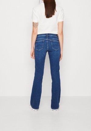 Femme aux longs cheveux bruns portant un jean bootcut bleu foncé, un t-shirt blanc à manches courtes, des baskets blanches et un tatouage toile d’araignée sur le haut du bras gauche.