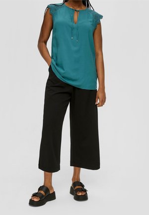 Vrouw draagt een turquoise mouwloze blouse met kanten schouders, losse zwarte korte broek en zwarte platformsandalen, staand met één hand in de zak.