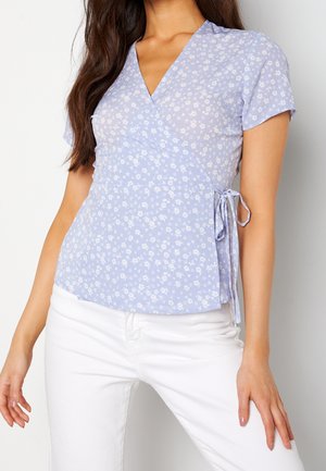 Femme portant un chemisier cache-cœur floral bleu clair à manches courtes et un pantalon blanc, posant avec une main sur la hanche.