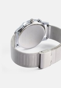 Silver mesh strap watch with a round stainless steel case, two crown buttons, and a smooth polished rear surface. Detailed engraving present.