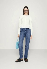 Blusa branca com pregas, jeans de denim azul, sandálias pretas estilo slide e uma pequena mala trançada azul, sobre um fundo neutro.