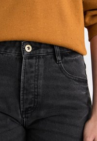 Jeans de mezclilla negros de cintura alta, con un botón y cremallera frontales visibles, exhibidos junto a un suéter texturizado mostaza.