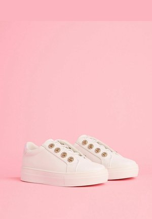 Fehér sportcipők vastag talppal, sima bőrből készült felső résszel és dekoratív aranyszínű gombokkal az egyik oldalon. Texturált gumitalp.