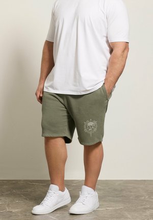 Homme portant une chemise blanche à manches courtes, un short vert olive avec un motif tête de mort et laurier, et des baskets blanches, debout sur un sol gris devant un mur beige.