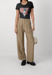 Žena v černém květovaném tričku s logem Guess, béžových širokých kalhotách, černých mokasínech a držící vedle sebe malou bílou kabelku.