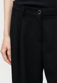 Detailný záber osoby nosiacej čierne nohavice s vysokým pásom so zapínaním na gombík a pútkami na opasok, ruka voľne cez bok.