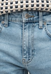 Κοντινή λήψη ανοιχτό μπλε τζιν παντελονιού με μεταλλικό κουμπί και θηλιές για ζώνη, συνδυασμένο με ένα μαύρο και λευκό πλεκτό τοπ.