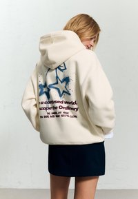 Kremowy hoodie z dużym graficznym nadrukiem niebieskich gwiazd i tekstu na plecach. Wykonany z miękkiego materiału, z luźnym krojem i kapturem.