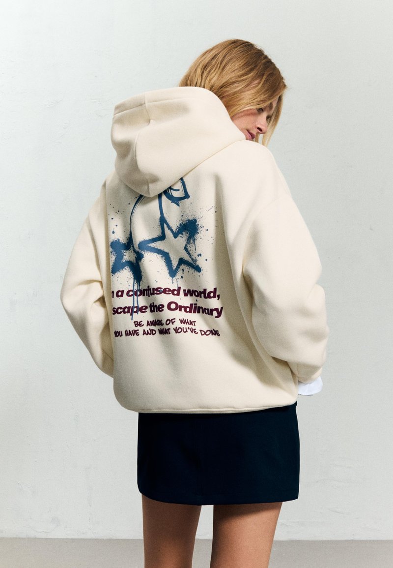 Kremowy hoodie z dużym graficznym nadrukiem niebieskich gwiazd i tekstu na plecach. Wykonany z miękkiego materiału, z luźnym krojem i kapturem.