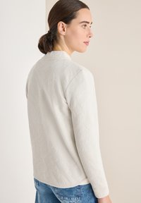 Světlý šedý svetr s texturovaným vzorem rybí kosti. Má kulatý výstřih a dlouhé rukávy, doplněný modrými džínami.