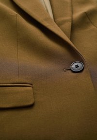 Hnědý blejzr z texturované tkaniny, s jediným černým knoflíkem, kapsou na levé straně a jemným detailem límce.