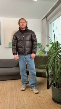 Black puffer jacket with a zip front, quilted design, and high collar, paired with light blue jeans and beige sneakers. Indoor setting with plants.