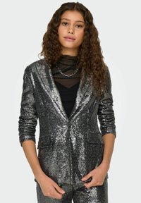 Blazer plateado con lentejuelas y mangas largas, con solapas de muesca y bolsillos delanteros. Llevado sobre una blusa negra, con una textura brillante.