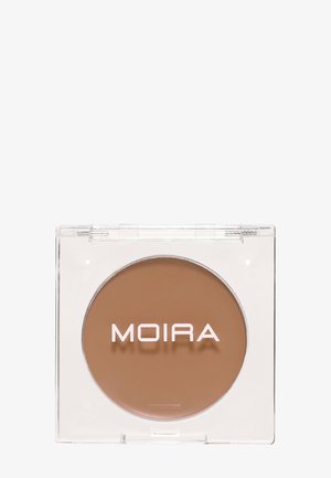 Pieni pyöreä ruskea voide meikkituote kirkkaassa neliönmuotoisessa muovikotelossa, jossa ylhäällä "MOIRA"-merkkitunnus.